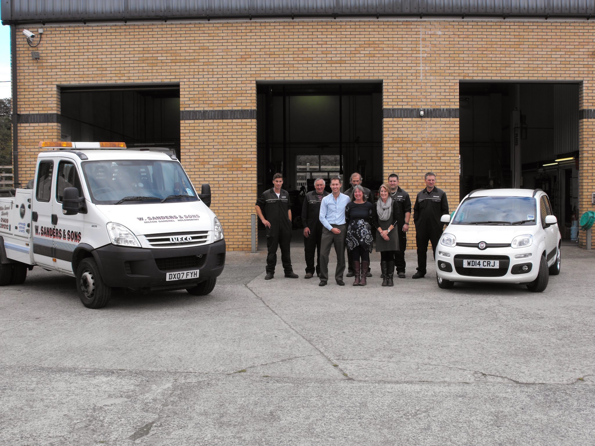 The W Sanders & Sons team lined up outside the workshop between the recovery truck and a Fiat Panda